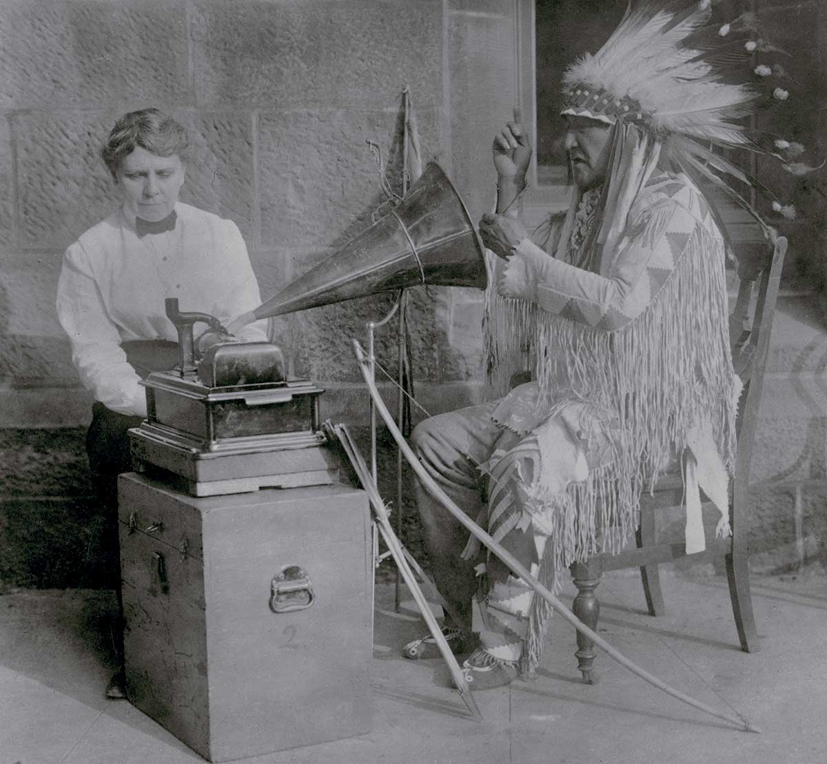 Archival black and white photograph of a man with a feathered headdress speaking into a recording device operated by a woman