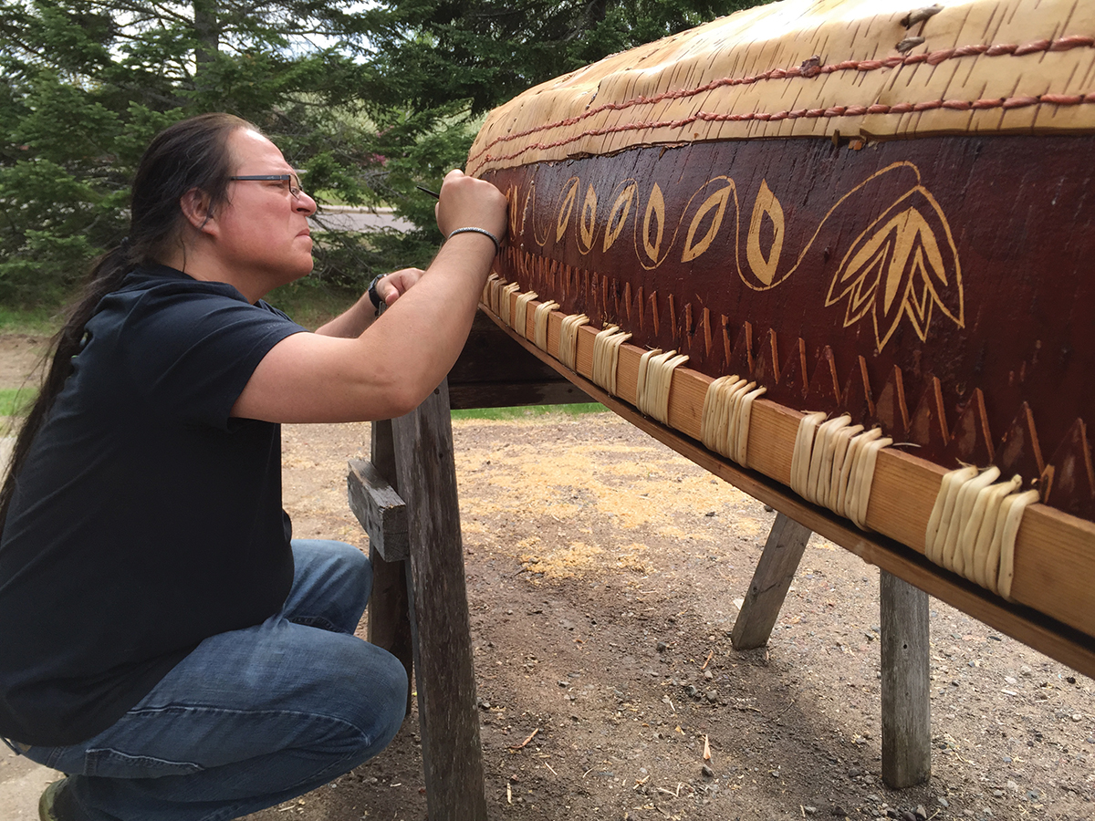 An artist crouches in front of a birchbark canoe, carving a design into its side