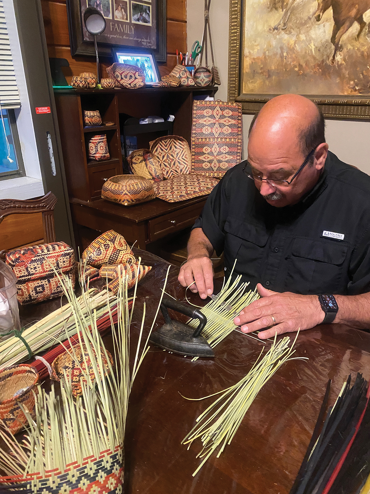 An artist seated at a table full of finished baskets weaves river cane into a basket