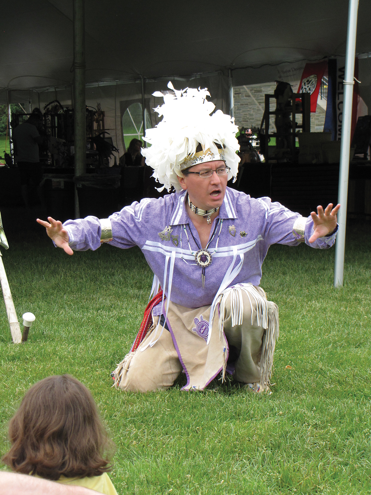 A storyteller in a headdress and purple shirt crouches during the telling of a story