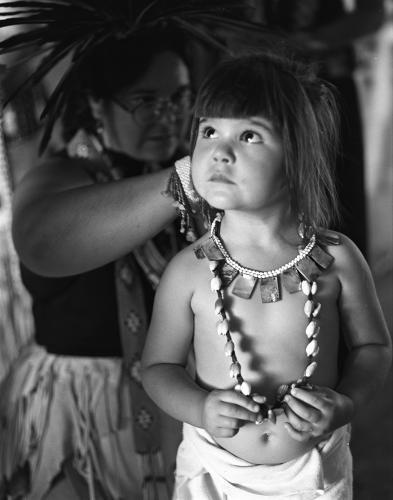 A black and white image. A mom puts necklaces on her young daughter for the dance.
