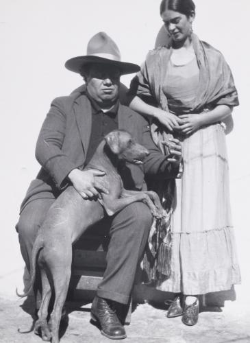 Diego Rivera sits in a chair with his medium-sized xoloitzcuintli dog propped up in his lap. Frida Kahlo stands to his right, looking at the dog.