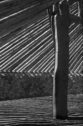 A black and white shot of a corner post from within a roundhouse. The wooden beams allow sunlight to stream into the space.