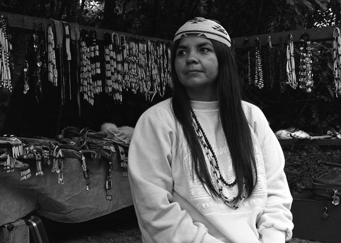 A black and white image of a woman standing in front of a stand filled with dance regalia. She looks off to the side.