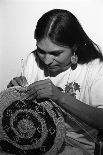 A black and white image of a woman, seated, hand-weaving a basket with a spiral design.