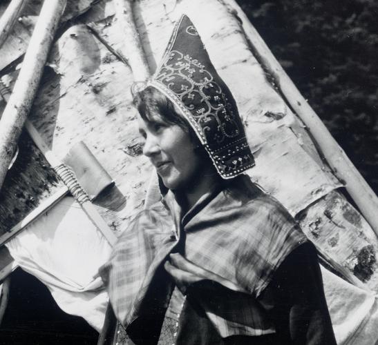 A black and white image of a woman wearing a peaked, embroidered cap and standing next to a cone-shaped dwelling