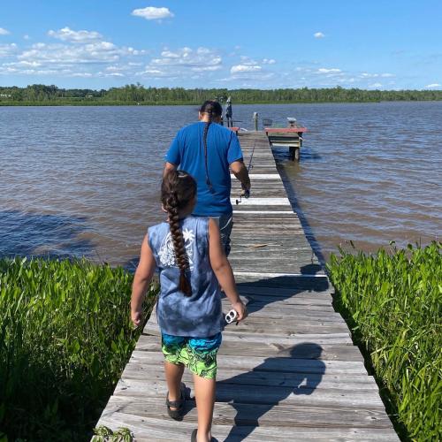 A father and son holding fishing rods walk down a dock toward a river