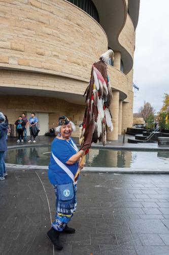 A woman dressed in blue regalia and wearing a U.S. Air Force cap carries a staff covered in eagle feathers