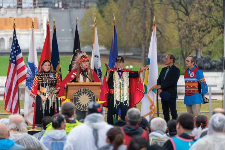 A man in a feather headdress stands at a podium