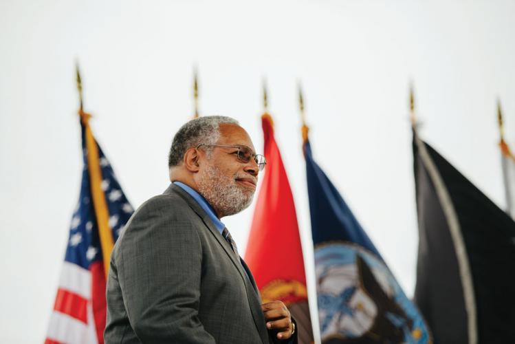A man wearing glasses and a grey suit is pictured in front of a series of flags