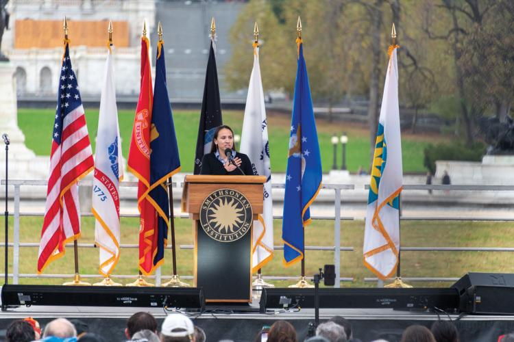 A woman stands at a podium in front of a variety of flags, holding her hand to her heart.