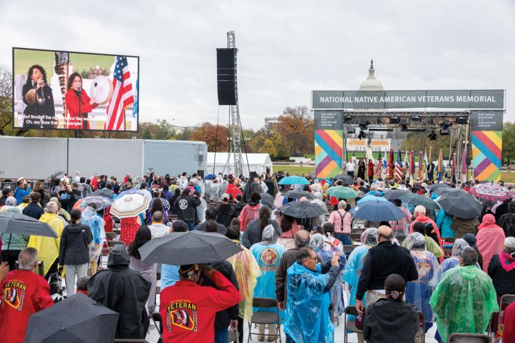 A crowd of people watches two musicians perform on an outdoor stage. The U.S. Capitol is visible in the background.