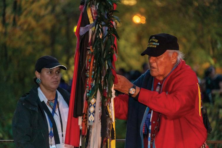 A man in a red jacket and Vietnam Veteran ball cap ties a cloth to a lance