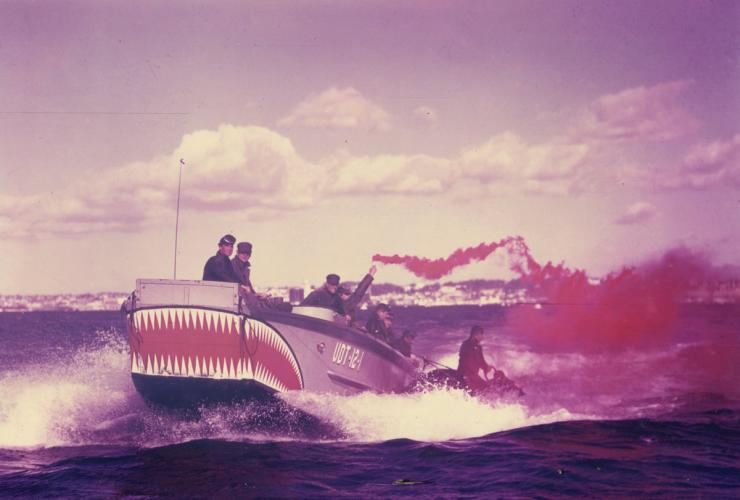 A 1950s photograph of a grey boat with teeth painted on one end carries 8 crew members on board. One crew member is holding a flare releasing red smoke.