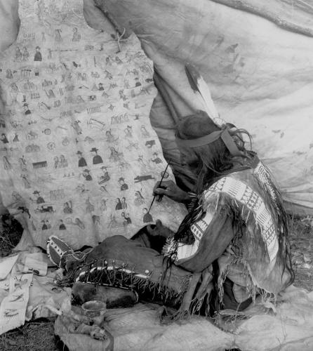 Black and white archival photo of a seated artist decorating a piece of hide