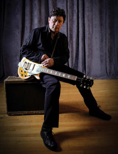 Robbie Robertson sits on an amplifier holding an electric guitar.