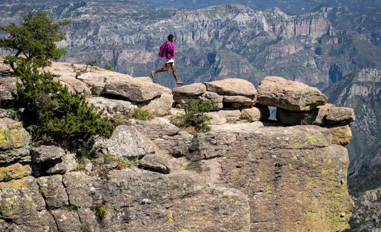 Person in traditional clothing and sandals running on top of rocky desert cliff.