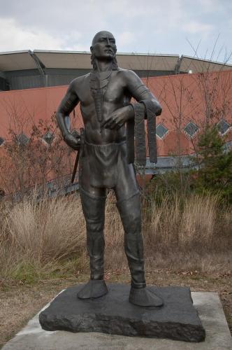 A bronze statue of Little Turtle stand tall in front of greenery.