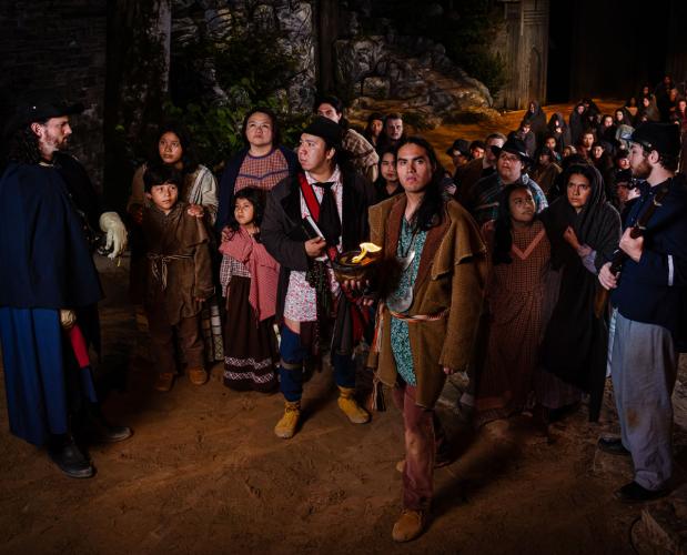 Actors on stage depict the Trail of Tears: a large group of Cherokee peoples are forcefully led by two soldiers.