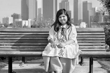 Black and white photograph of a young girl in Yalálag clothing, sitting on a park bench with the Los Angeles skyline behind her